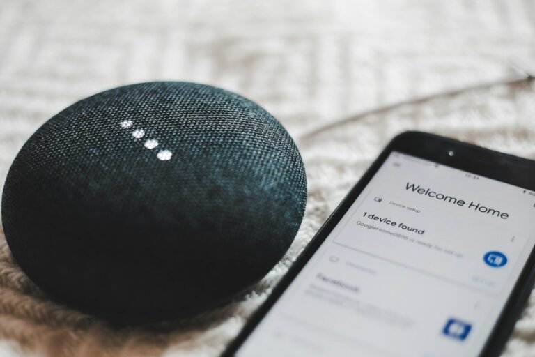 A charcoal Google Home Mini smart speaker next to a smartphone showing a smart home app